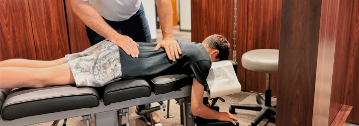 A chiropractic adjustment in progress, with a person lying face down on a treatment table while the practitioner applies pressure to the back.