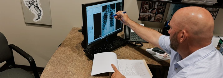 Dr. Quinn Uzelman reviewing and pointing at chest X-ray images displayed on a computer screen, with notes and a phone on the desk.