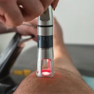 A close-up of a handheld device emitting red light being applied to a person’s knee, likely for therapeutic treatment.