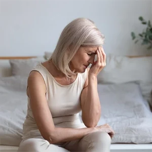 An individual seated on a bed, resting their head on one hand, suggesting discomfort or fatigue.